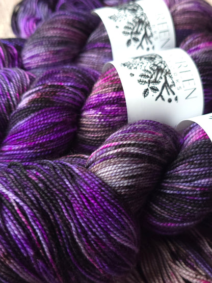 Close-up of several skeins of variegated yarn in deep purple, violet, and black tones, with white paper labels wrapped around some skeins. The brand name appears on the labels.