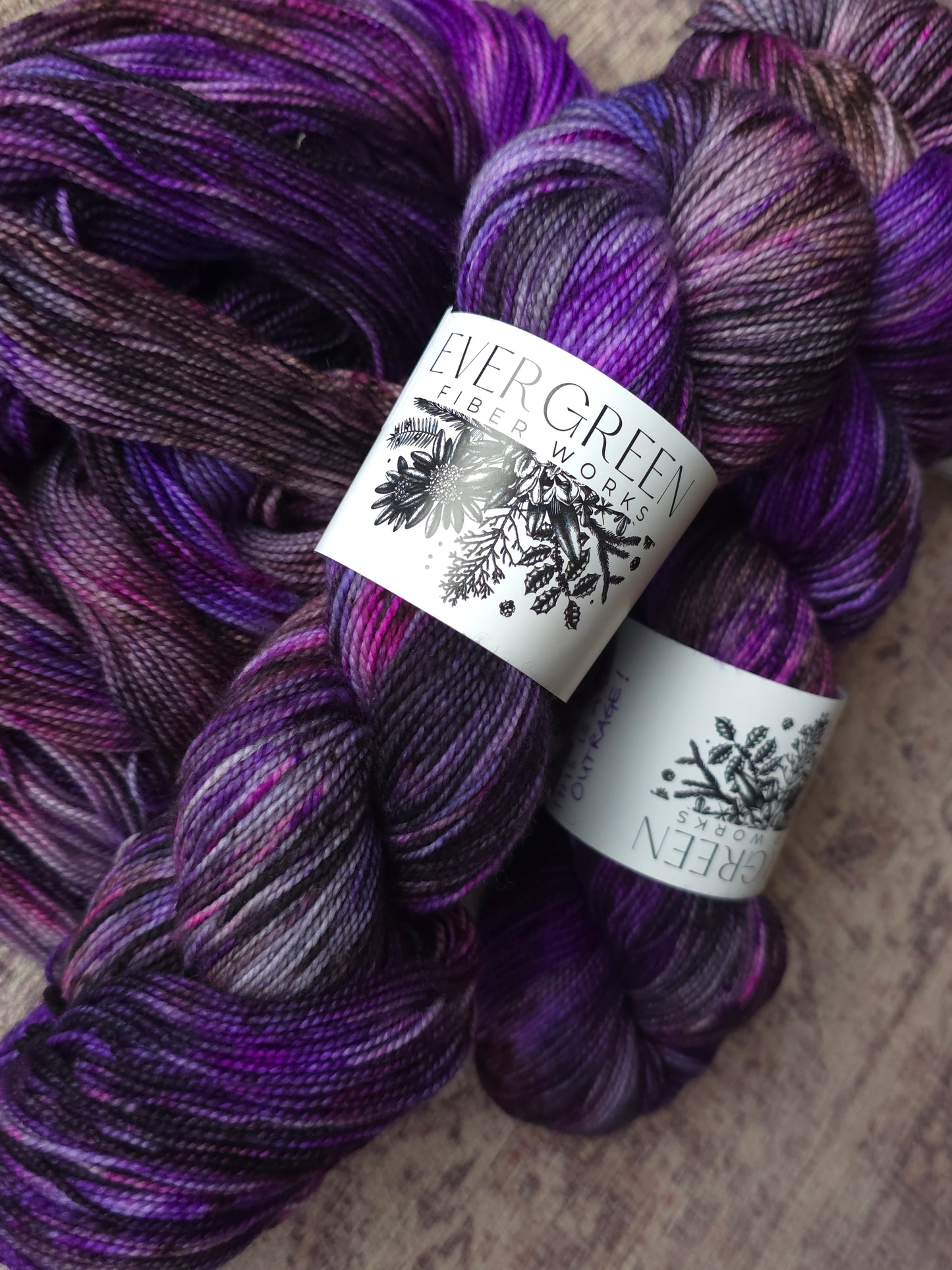 Two skeins of Evergreen Fibre Yarn in a variegated colorway, showing a blend of deep purple, violet, and dark gray tones, with white paper labels featuring black botanical illustrations.